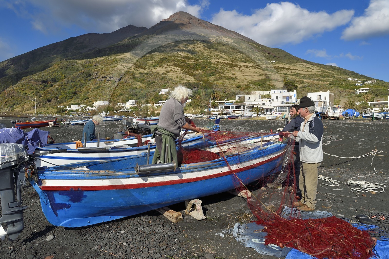 Italy, Sicily, Aeolian Islands, listed as World Heritage by UNESCO, Stromboli island, the fisherman Gaetano Cusolito repairing nets on the beach of Scari with his two brothers and the volcano Stromboli in the background