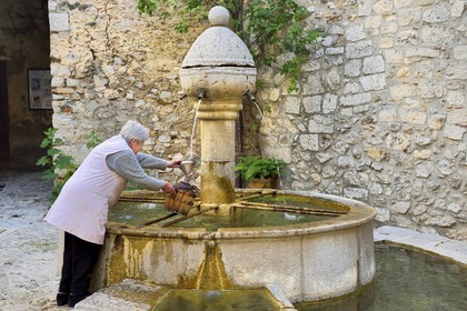 France, Alpes-Maritimes, Peille, woman filling a bottle at the fountain place du Mont Agel